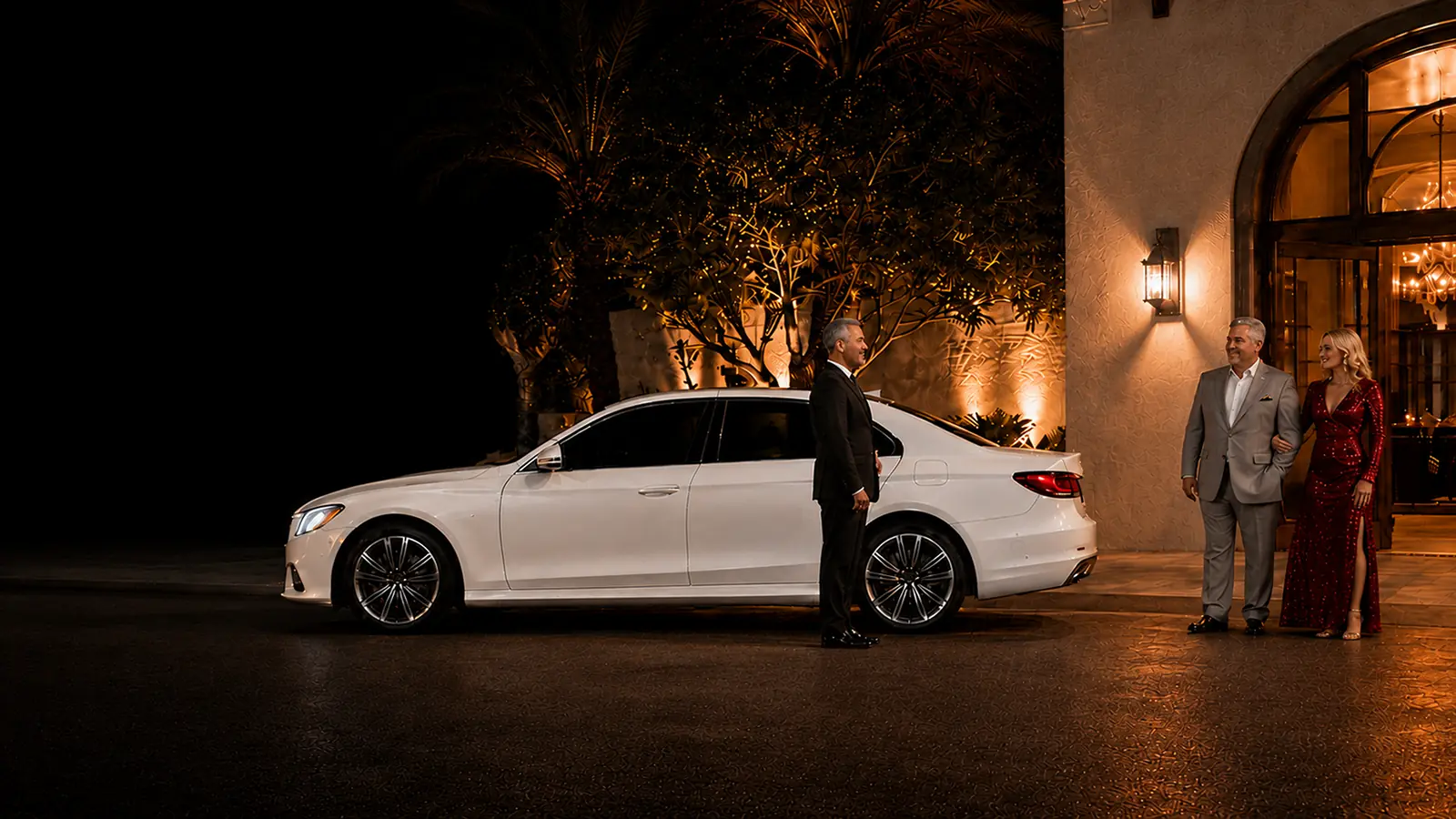 Private driver greeting a couple beside their white sedan outside a restaurant.