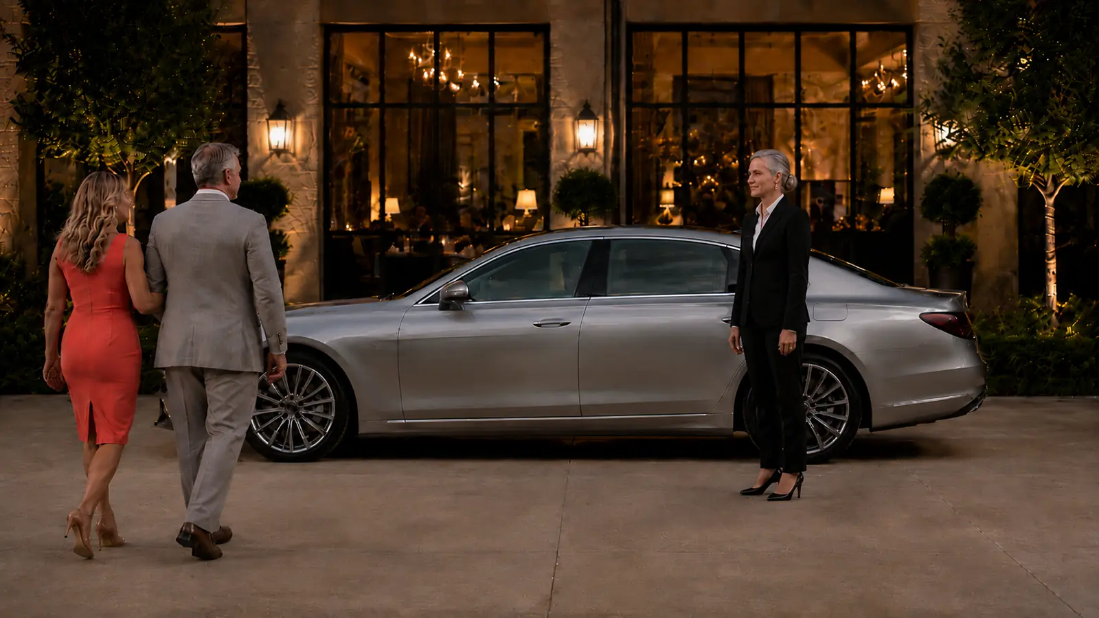 Private driver waiting beside a silver sedan as a couple leaves a restaurant in late afternoon.