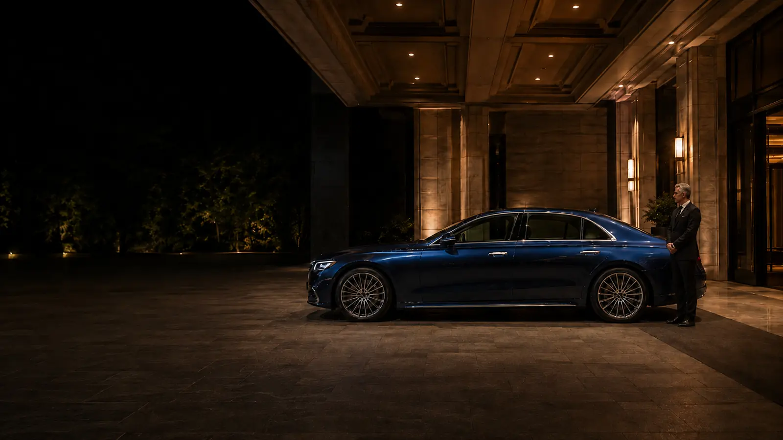 Private driver waiting beside a client's luxury blue sedan at an upscale residence at night.